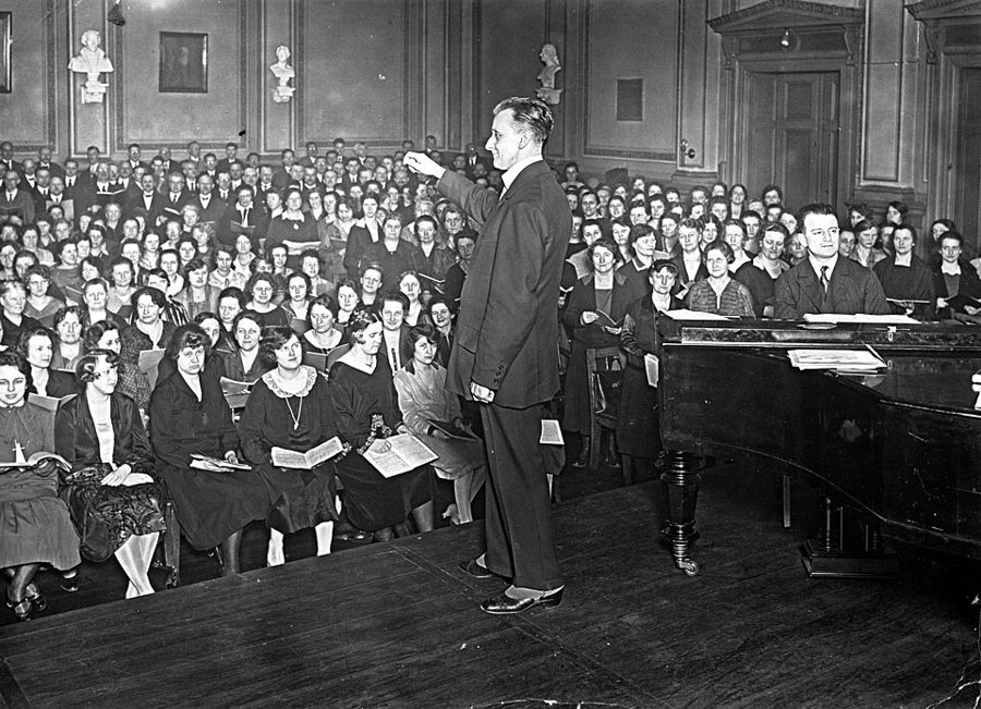 Dyrygent stoi przed dużym chórem w sali, obok pianista przy fortepianie